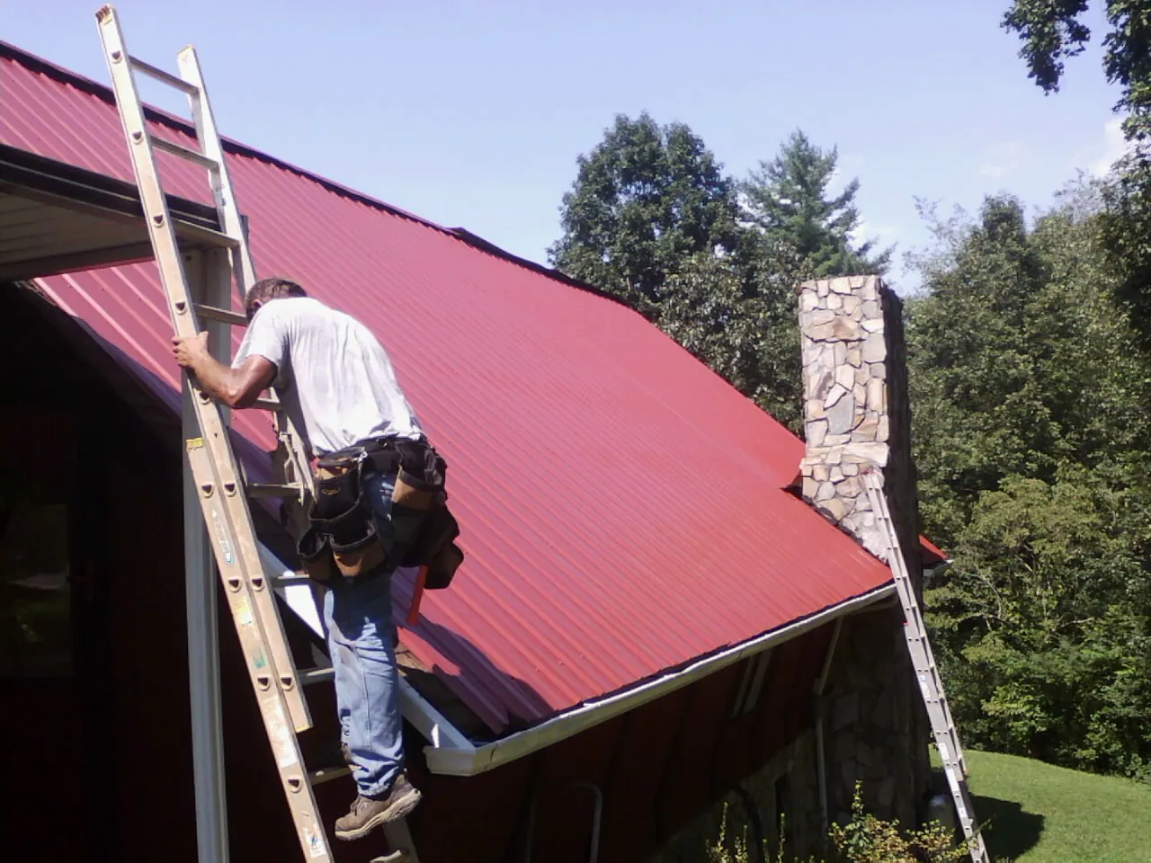Roofer on a ladder beside a red metal roof and stone chimney — Soffit & Fascia Repair by Roofing Brothers in Manheim